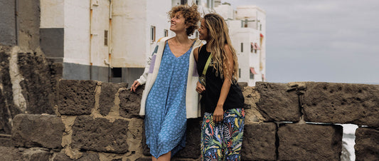 Two women wearing summer clothes lean against a wall smiling