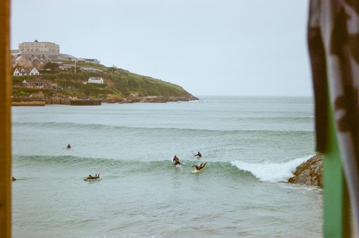 A scenic coastal view with surfers floating on boards in the ocean, a rocky shoreline, and buildings on a grassy cliff under a pale sky. The photo is framed by blurred vertical foreground elements on both sides.